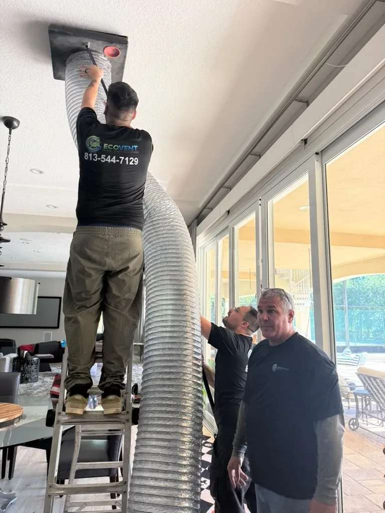 Un técnico de Ecovent, subido a una escalera de tijera, conecta una manguera ancha de ducto flexible a una rejilla del techo de una sala mientras dos compañeros esperan abajo y uno sujeta la manguera