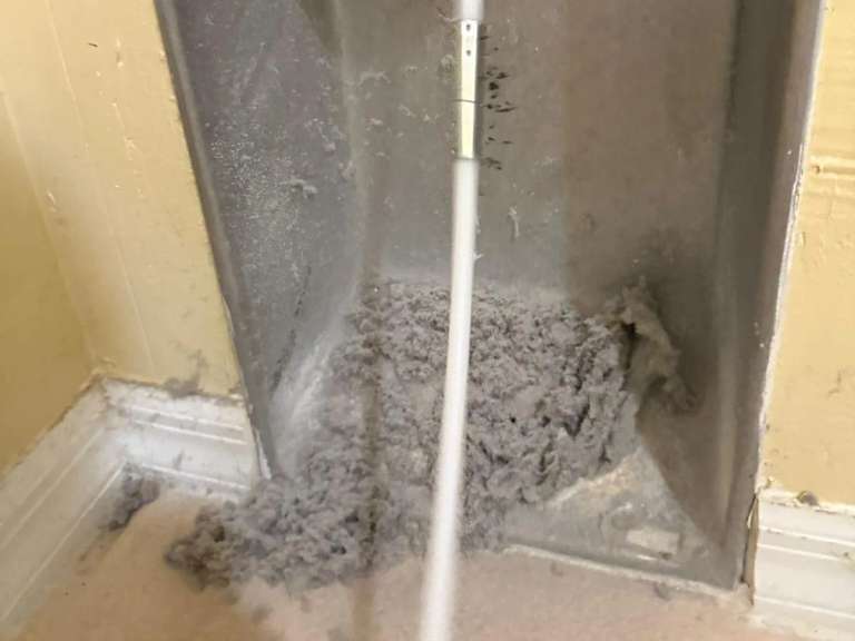 Pelusa amontonada al fondo de la caja de pared de un ducto de secadora, con el cable del cepillo de limpieza subiendo por el ducto