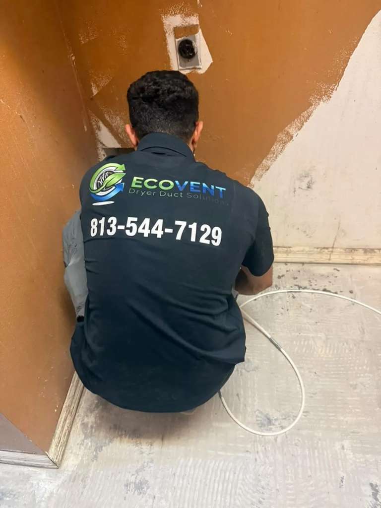 An Ecovent technician crouched at a dryer vent wall connection in a laundry room, the cleaning cable coiled on the floor beside him