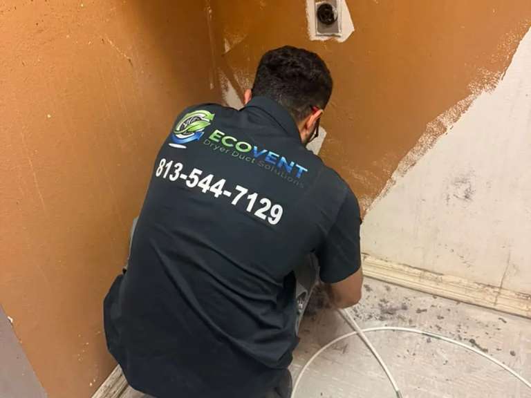 Un técnico de Ecovent arrodillado junto a la conexión de pared de un ducto de secadora, metiendo el cable del cepillo en el ducto, con pelusa en el piso