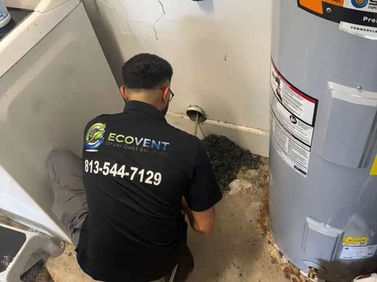An Ecovent technician kneeling between a washer and a water heater clears a dryer vent at the wall connection, with the lint he pulled out piled on the floor