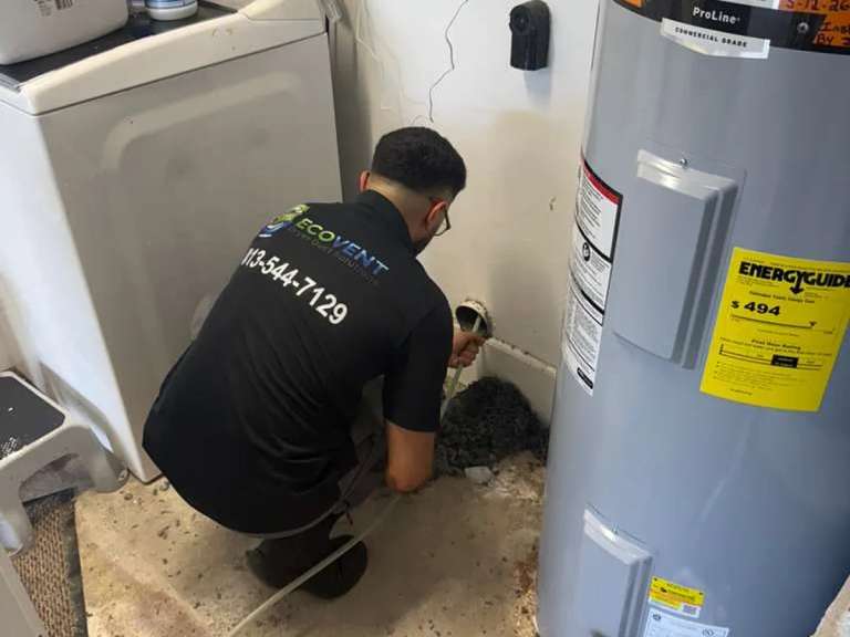 Un técnico de Ecovent agachado en un clóset de lavado, entre la lavadora y el calentador de agua, trabajando en la conexión del ducto de la secadora
